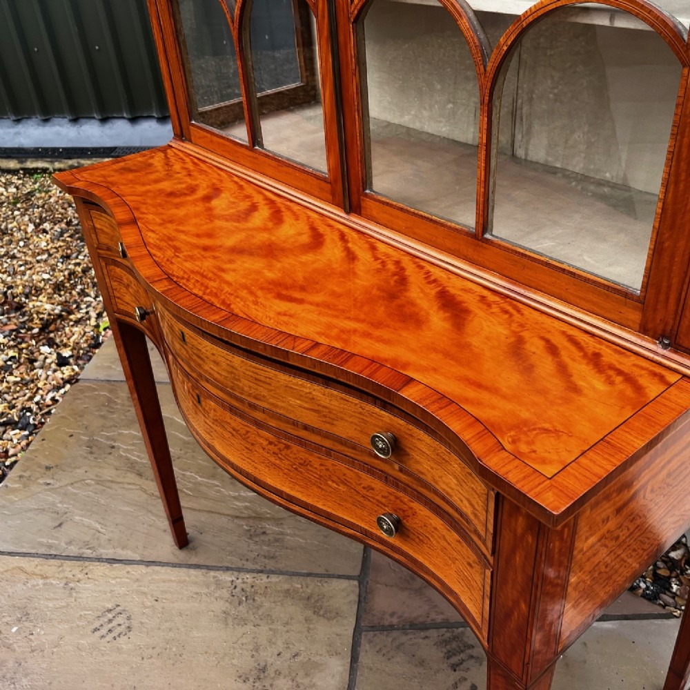 An antique Sheraton Revival satinwood display cabinet featuring arched astragal glazed doors, two lower drawers with ring handles, and square tapered legs. View IV