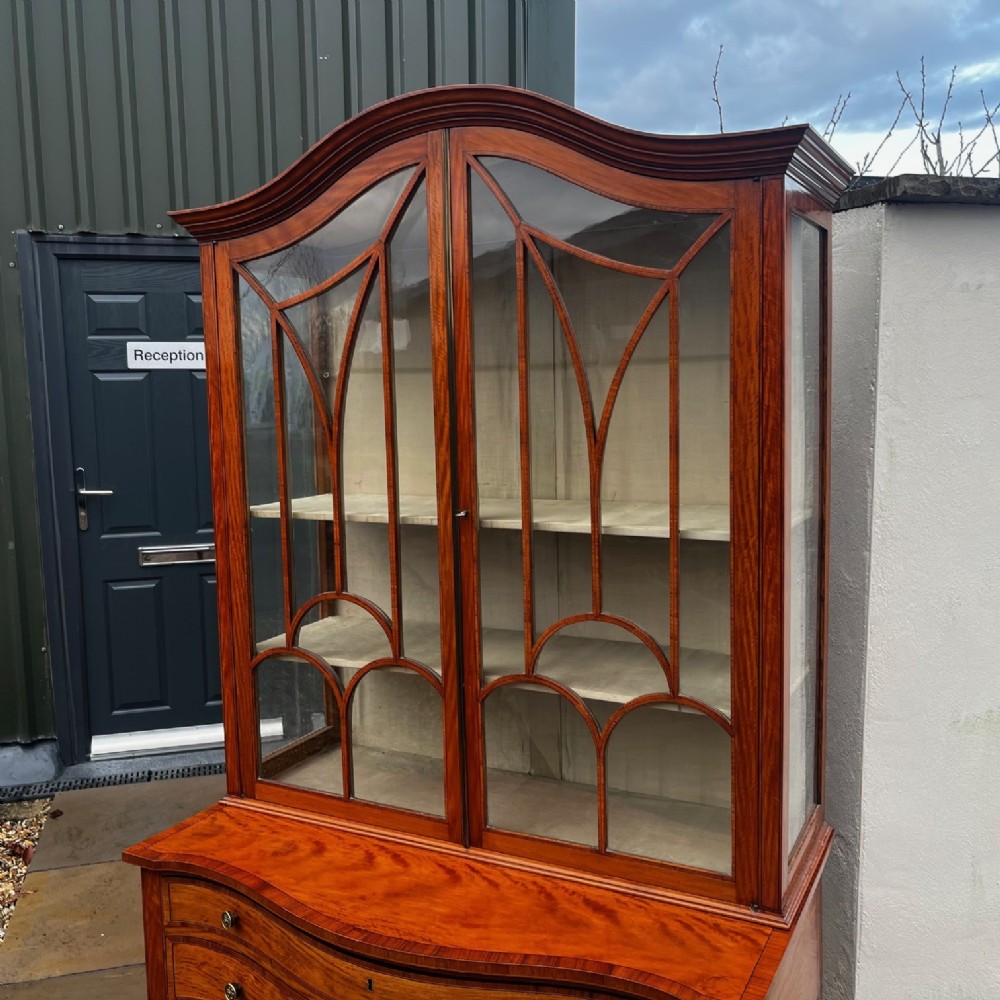 An antique Sheraton Revival satinwood display cabinet featuring arched astragal glazed doors, two lower drawers with ring handles, and square tapered legs. View V