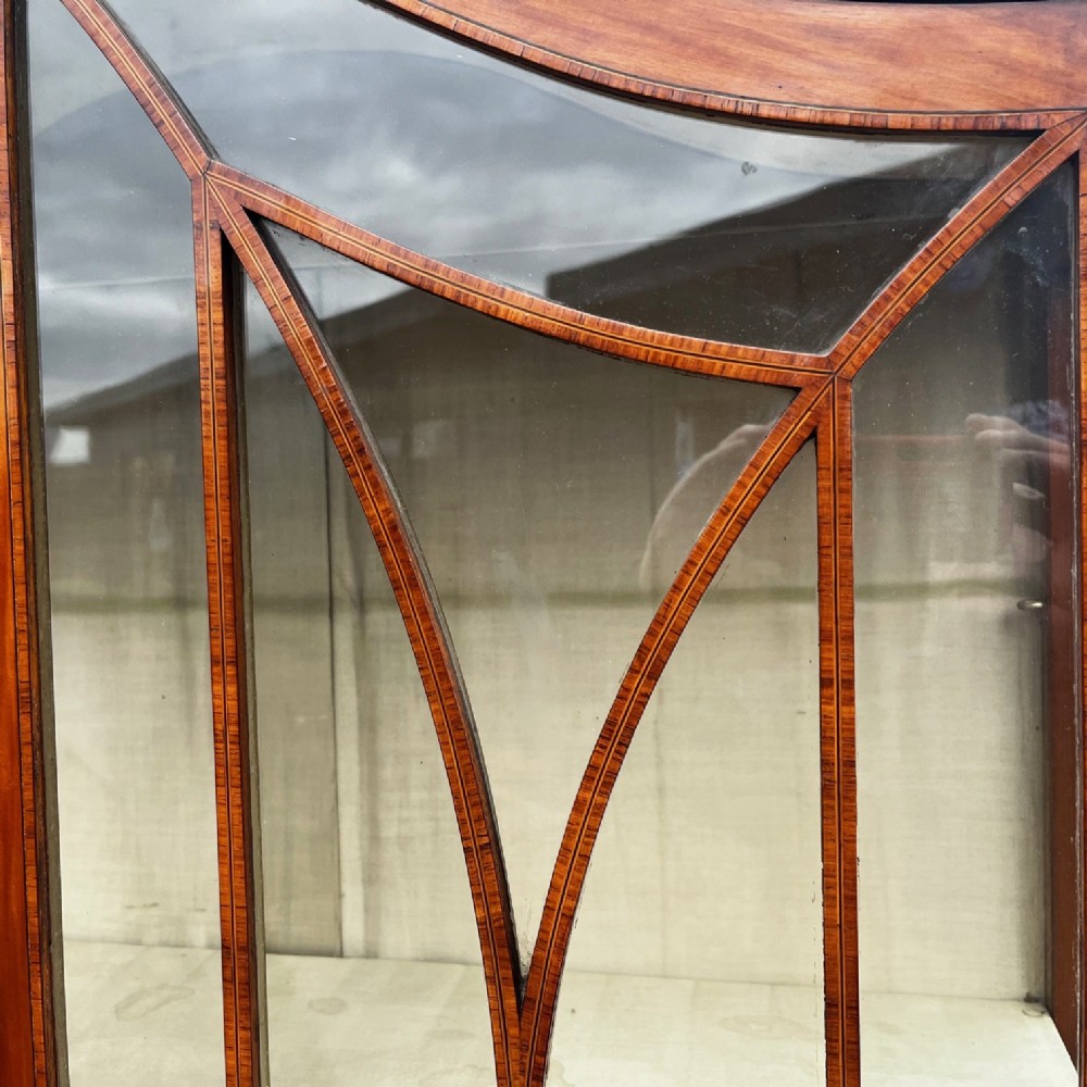An antique Sheraton Revival satinwood display cabinet featuring arched astragal glazed doors, two lower drawers with ring handles, and square tapered legs. View VI
