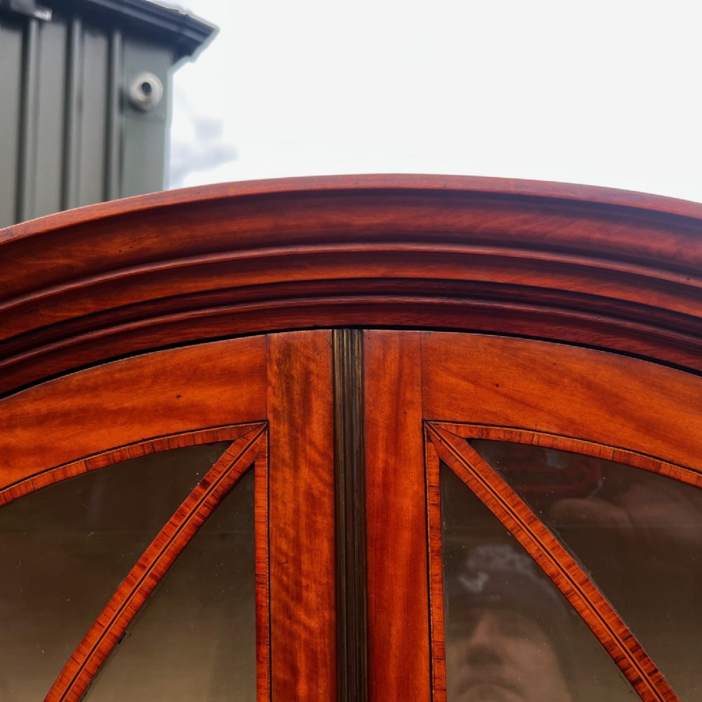 An antique Sheraton Revival satinwood display cabinet featuring arched astragal glazed doors, two lower drawers with ring handles, and square tapered legs. View VII