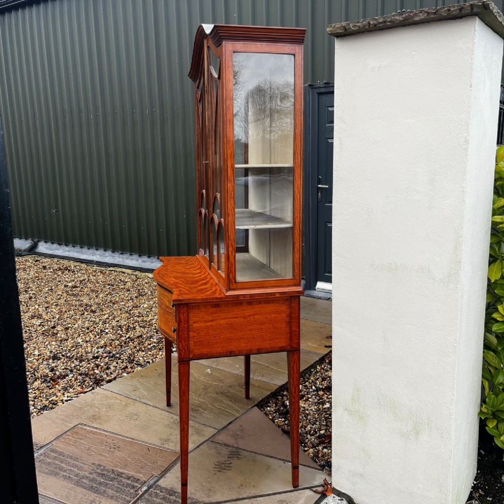 An antique Sheraton Revival satinwood display cabinet featuring arched astragal glazed doors, two lower drawers with ring handles, and square tapered legs. View VIII