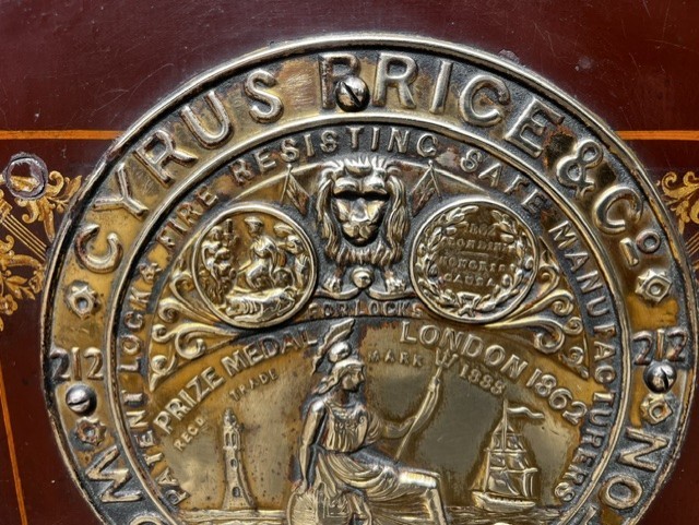 A Victorian cast iron safe featuring a black outer body, maroon door with gold pinstriping, and a round brass Cyrus Price & Co maker badge. View III