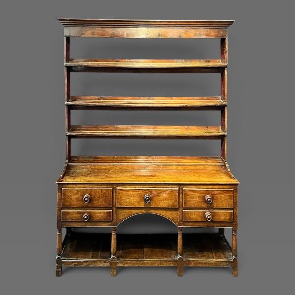 An early 19th-century dark oak Welsh dresser featuring a tall three-shelf open plate rack mounted on a base with five drawers, turned legs, and a lower pot board shelf.