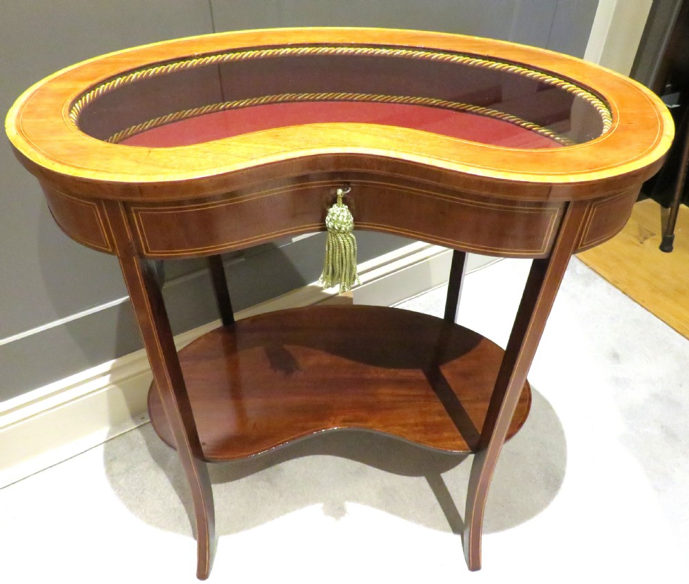 An Edwardian kidney-shaped mahogany bijouterie table featuring fine satinwood inlay, a glass-topped display section lined in burgundy fabric, and a lower shelf.