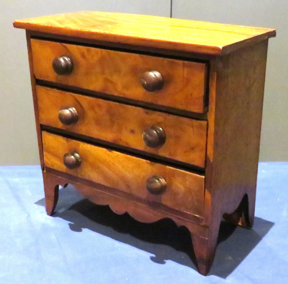 A wooden miniature chest featuring three drawers with turned knobs and a shaped lower apron.