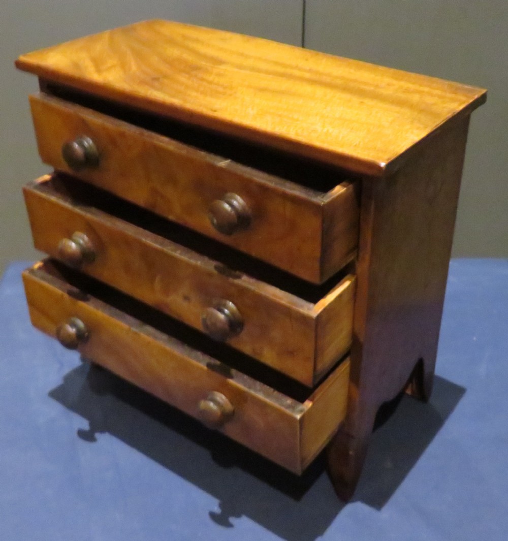 A wooden miniature chest featuring three drawers with turned knobs and a shaped lower apron. View II