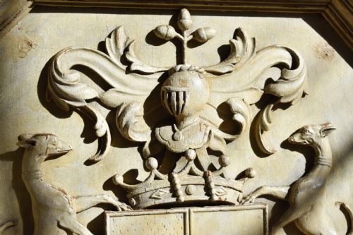fig 1. An octagonal framed timber plaque featuring a high-relief carved coat of arms with hounds, a helmet, and a Latin motto scroll, finished in distressed off-white paint.