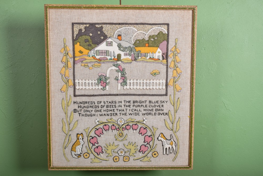 A framed textile needlework picture featuring a stitched country cottage landscape, a four-line poem, foxgloves, bleeding hearts, a cat, and a dog on a natural linen ground.