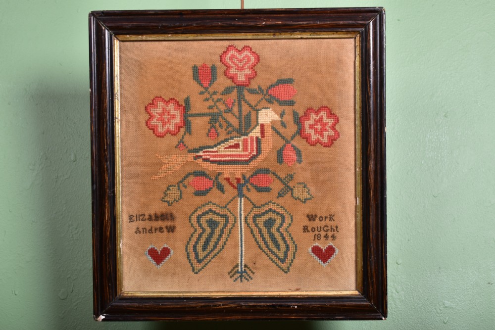 An 1844 Victorian needlework sampler framed in dark wood featuring a central bird, red flowers, two hearts, and hand-stitched lettering.