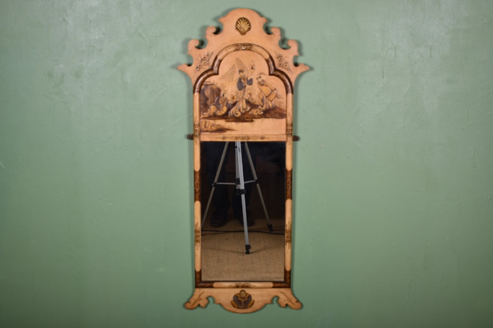 A tall, narrow cream painted wall mirror featuring a shaped scroll crest with a central shell motif and a hand-painted chinoiserie scene above the rectangular glass.