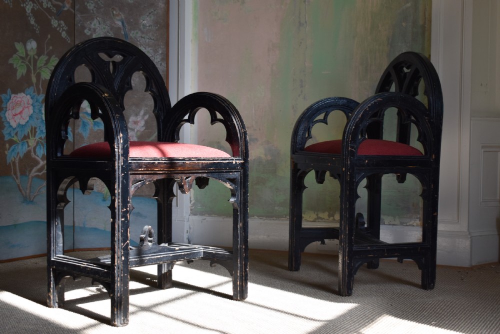 A pair of distressed black painted Gothic Revival armchairs featuring carved lancet tracery cutouts and red upholstered seats.