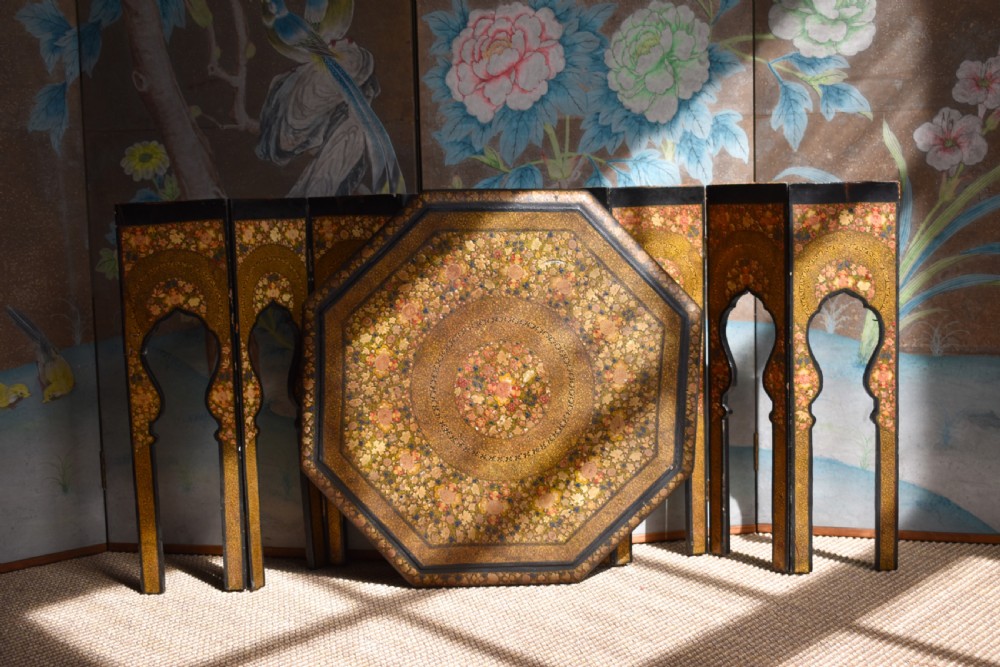 An octagonal Indian Kashmiri side table featuring intricate hand-painted floral motifs in gold, red, and green on a dark lacquered ground, resting alongside its matching folding arch-shaped base.
