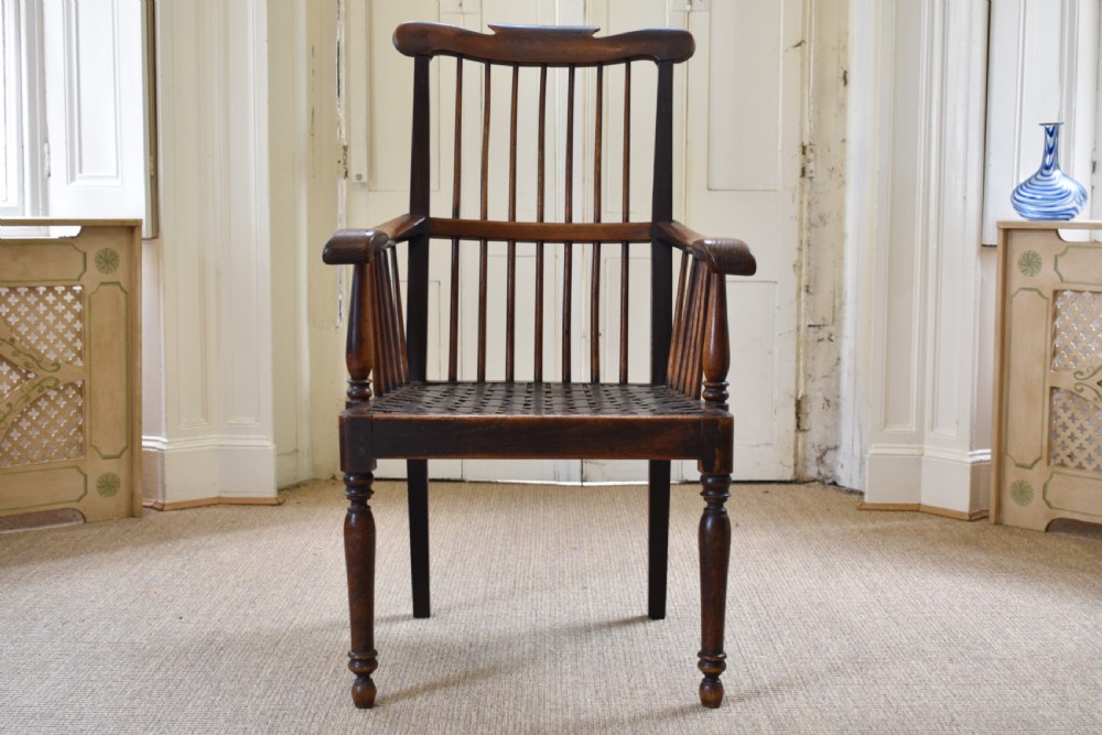 A dark timber country armchair featuring a tall spindle backrest, flat armrests on turned supports, a woven seat, and turned front legs.