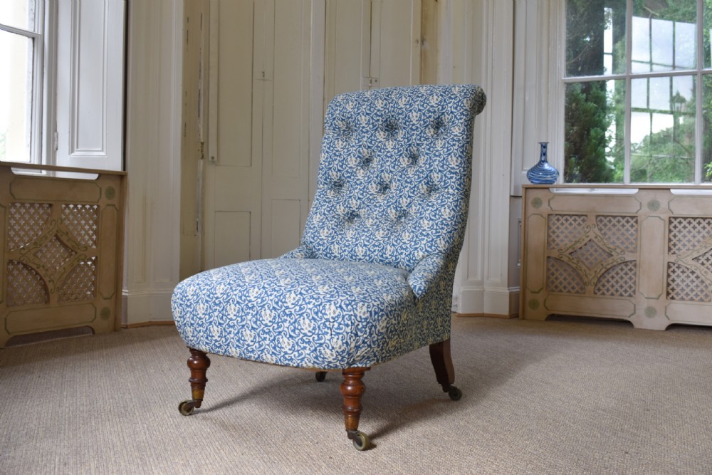 A low-seated Victorian slipper chair upholstered in blue and white patterned fabric with a button-tufted back and turned mahogany front legs on brass castors.