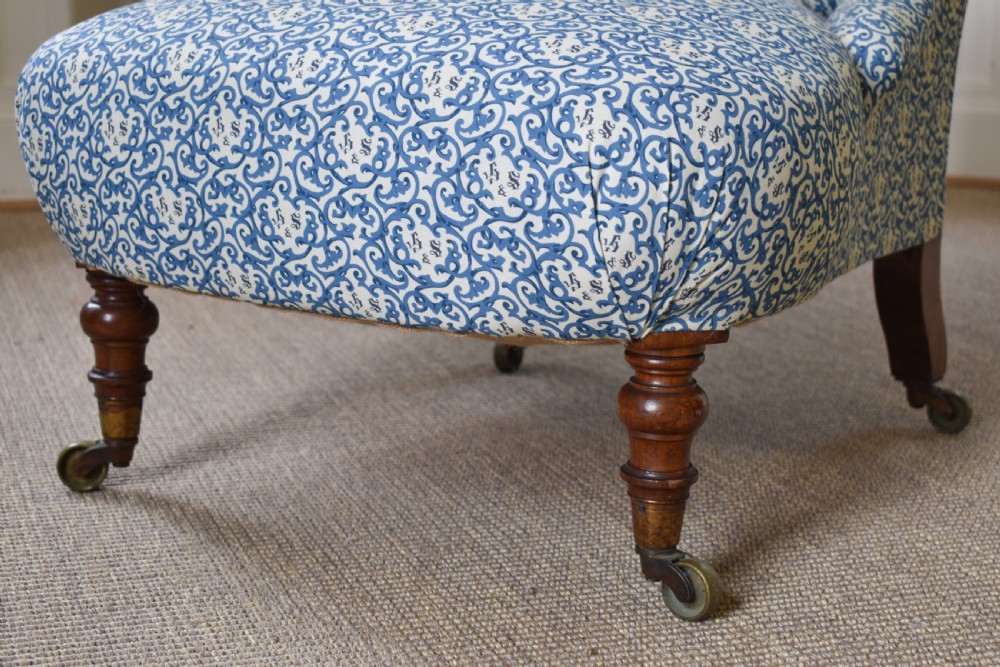 A low-seated Victorian slipper chair upholstered in blue and white patterned fabric with a button-tufted back and turned mahogany front legs on brass castors. View II