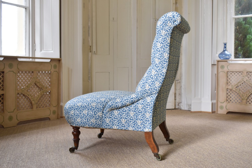 A low-seated Victorian slipper chair upholstered in blue and white patterned fabric with a button-tufted back and turned mahogany front legs on brass castors. View V