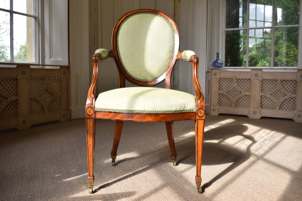A George III satinwood open armchair featuring an oval upholstered backrest, light green fabric, padded armrests, and tapered legs on brass castors.