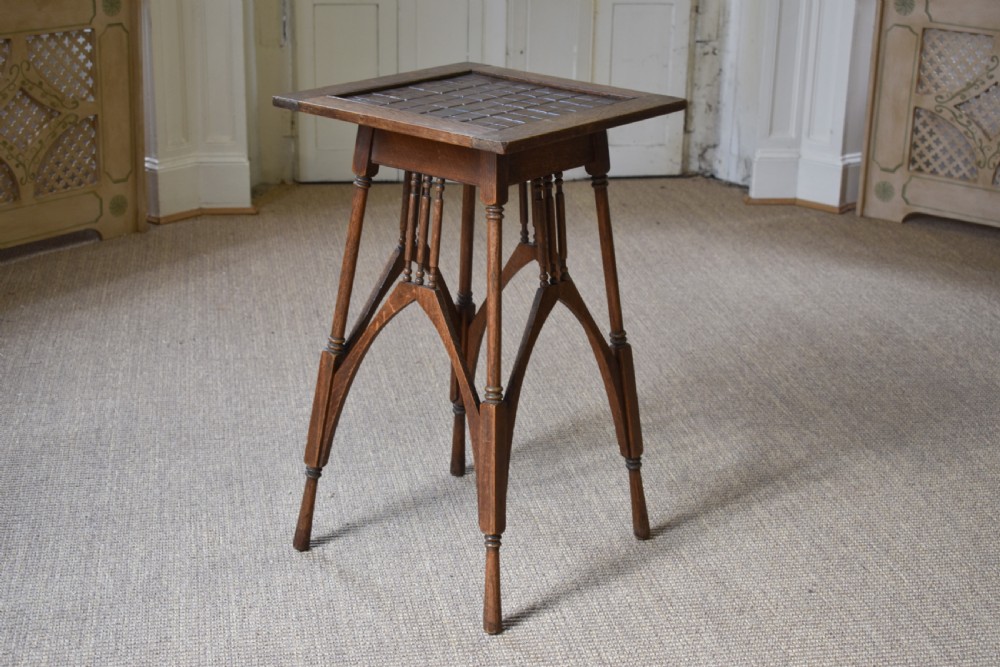 An antique Arts and Crafts oak occasional table featuring a square tiled inset top, arched bracing, and slender turned spindle legs.