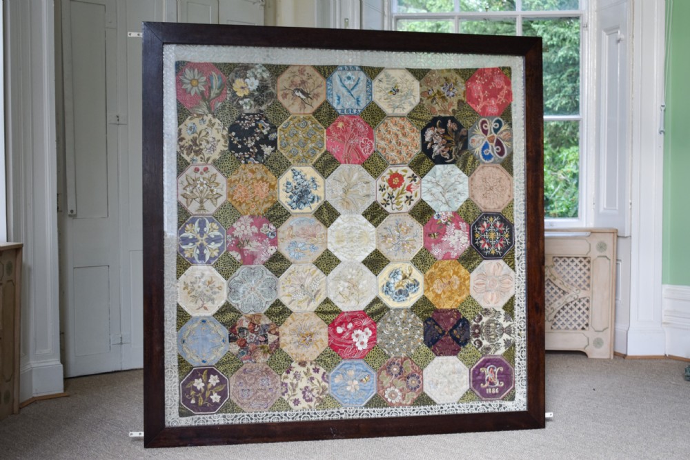 A large square framed Victorian silk patchwork panel featuring octagonal segments with intricate embroidered floral motifs and a dated section in the corner.