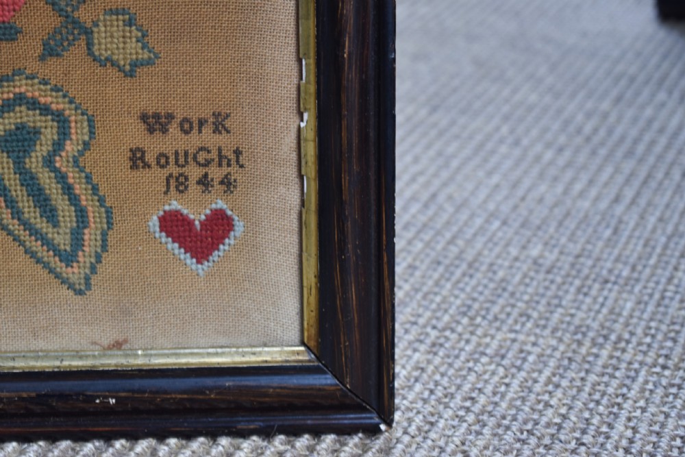 A framed 1844 Victorian needlework sampler featuring an embroidered bird perched on stylized floral stems with hearts and lettering, set in a dark wood frame. View IV