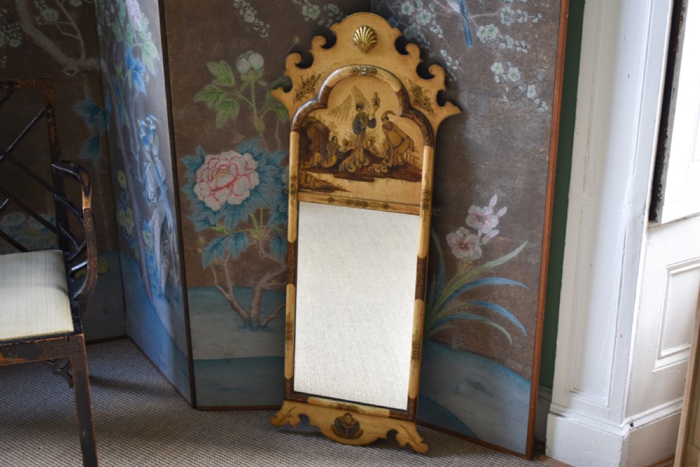 A tall, narrow Queen Anne style mirror featuring an arched frame decorated with chinoiserie hand-painted scenes, a carved gilt shell cresting, and ochre yellow lacquered borders.
