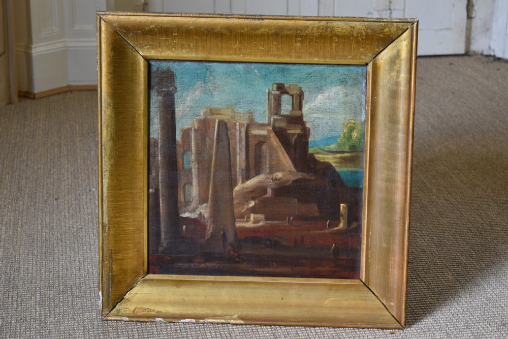 An antique oil painting depicting classical architectural ruins with columns and an obelisk set within a wide gold gilt frame.