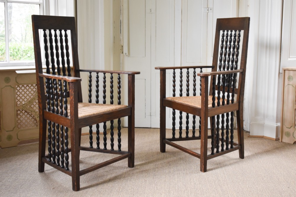 A pair of dark oak high-backed armchairs featuring bobbin-turned spindle backs, open arms with spindle supports, and woven rush seats. View III