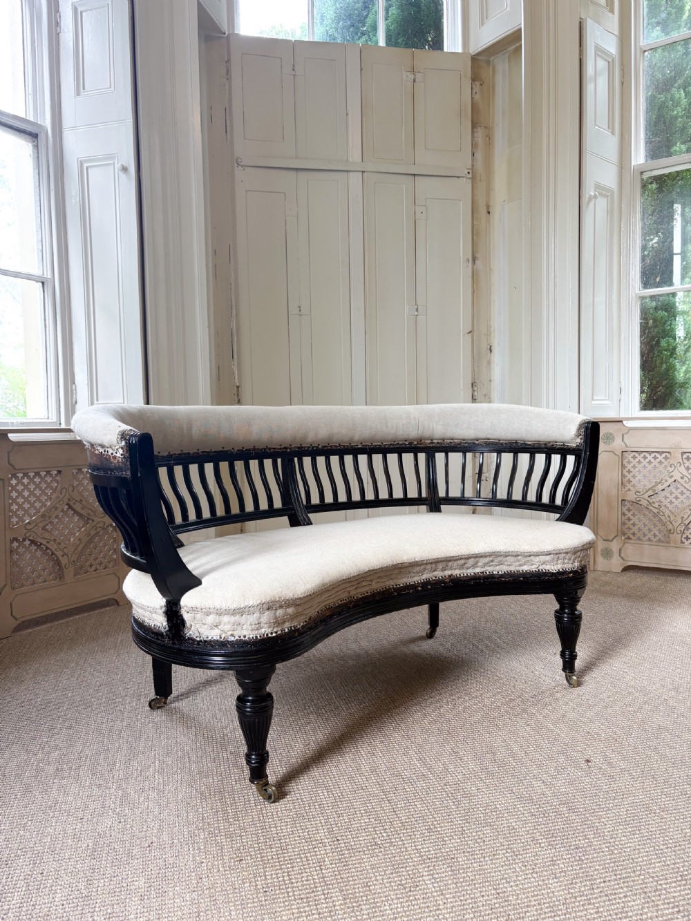 A Victorian ebonised curved settee featuring a spindle backrest, neutral calico upholstery, turned legs, and brass castors.