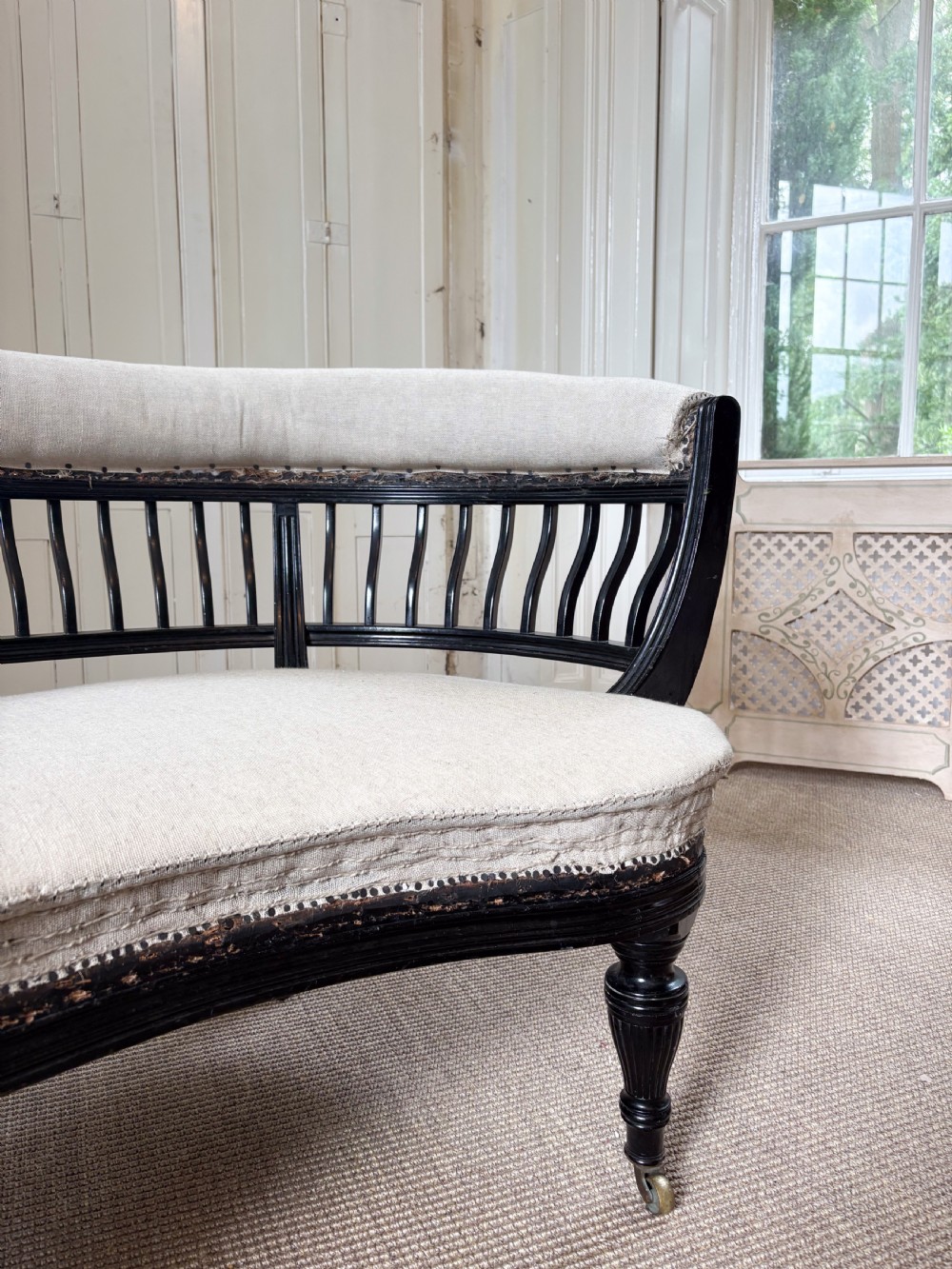 A Victorian ebonised curved settee featuring a spindle backrest, neutral calico upholstery, turned legs, and brass castors. View II