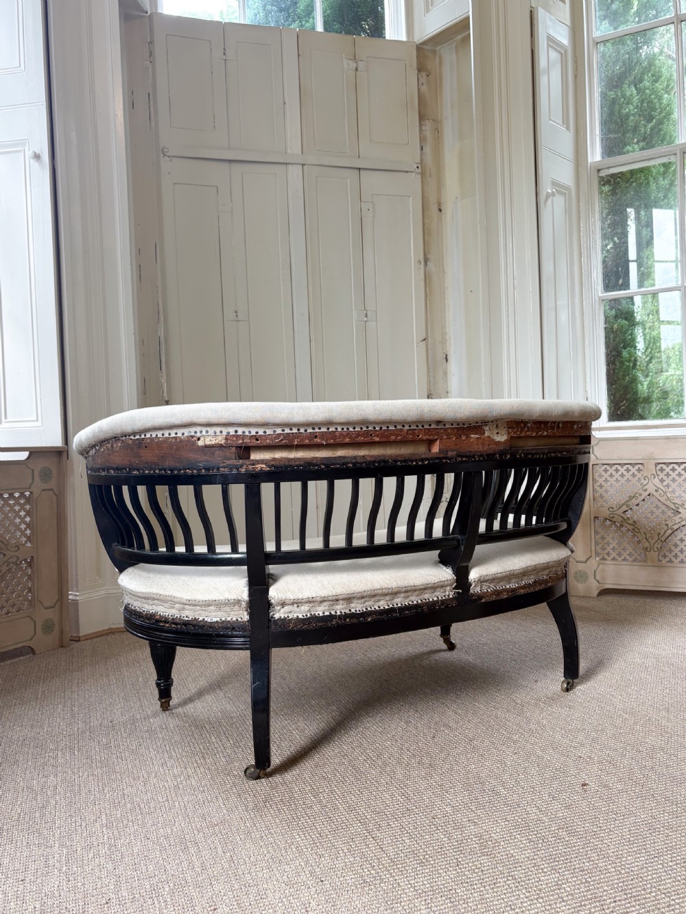 A Victorian ebonised curved settee featuring a spindle backrest, neutral calico upholstery, turned legs, and brass castors. View III