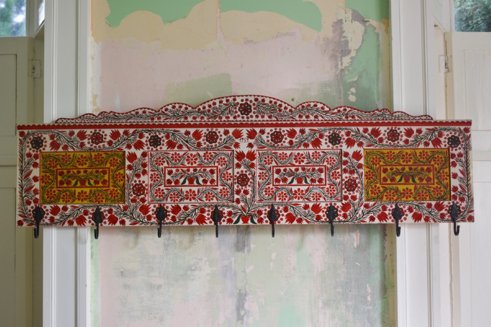 A long wall-mounted wooden coat rack featuring hand-painted red and yellow floral folk art patterns, a scalloped top edge, and eight iron hooks.