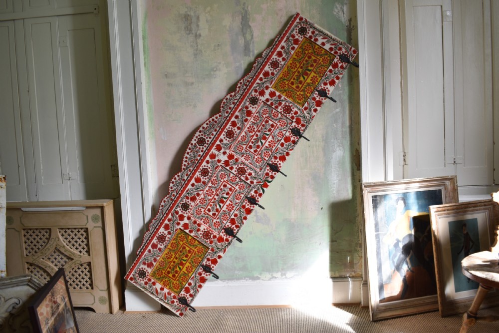 A long wall-mounted wooden coat rack featuring hand-painted red and yellow floral folk art patterns, a scalloped top edge, and eight iron hooks. View IV