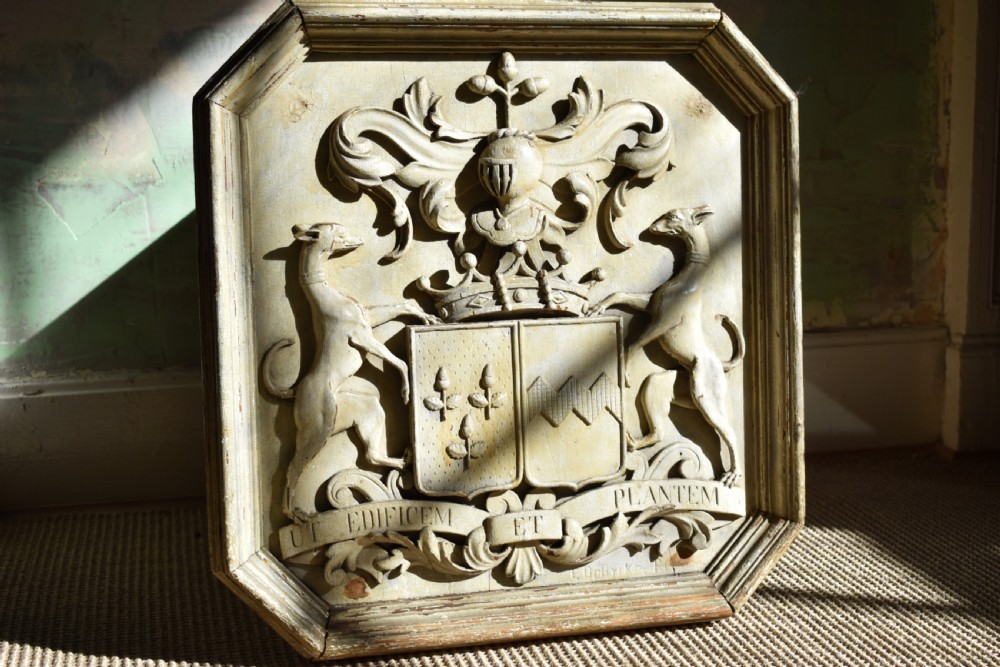 An octagonal framed timber plaque featuring a high-relief carved coat of arms with hounds, a helmet, and a Latin motto scroll, finished in distressed off-white paint.