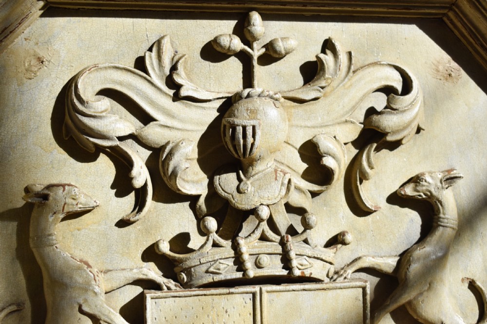 An octagonal framed timber plaque featuring a high-relief carved coat of arms with hounds, a helmet, and a Latin motto scroll, finished in distressed off-white paint. View II