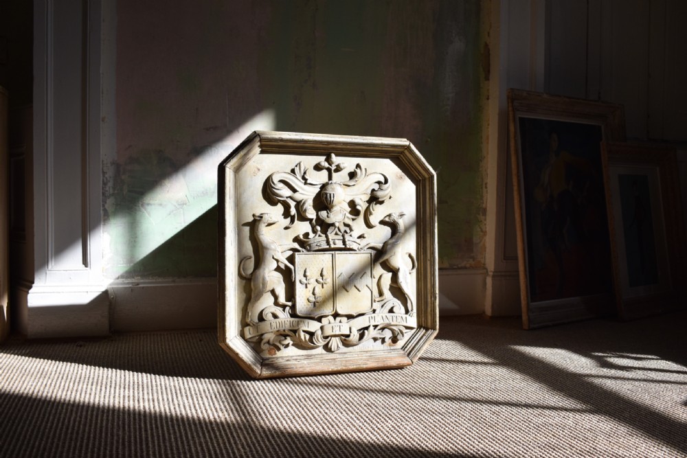 An octagonal framed timber plaque featuring a high-relief carved coat of arms with hounds, a helmet, and a Latin motto scroll, finished in distressed off-white paint. View III