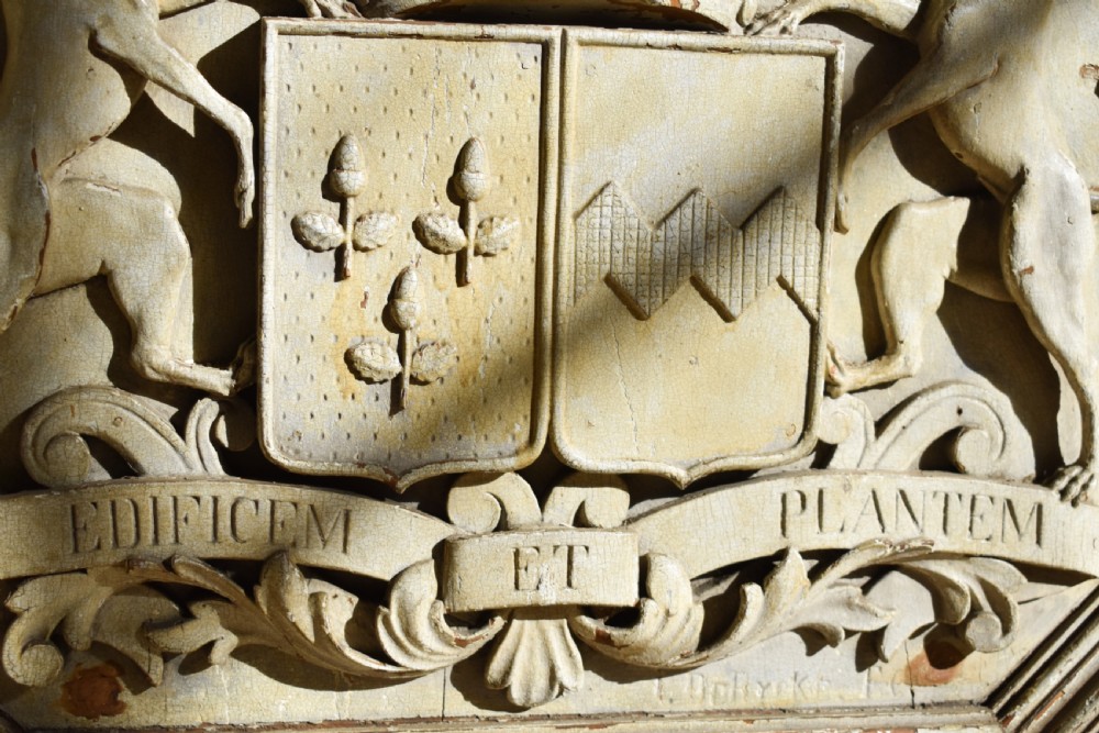 An octagonal framed timber plaque featuring a high-relief carved coat of arms with hounds, a helmet, and a Latin motto scroll, finished in distressed off-white paint. View IV