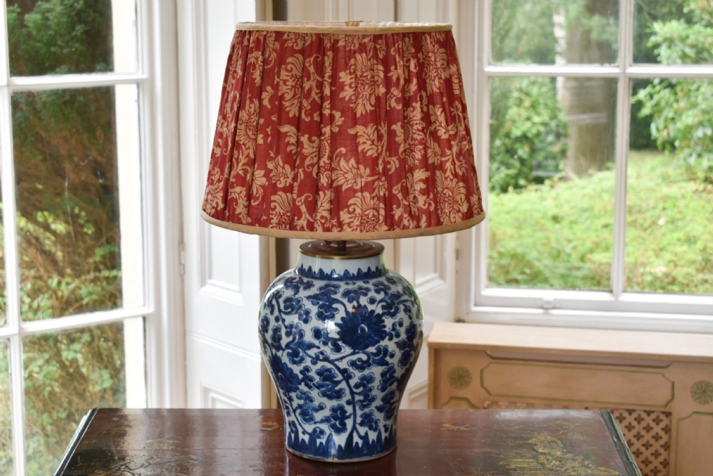 A Chinese blue and white hand-painted porcelain vase adapted as a table lamp with a pleated red floral patterned shade.