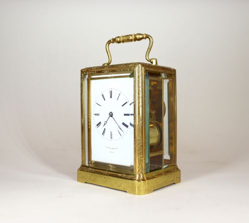 fig 1. An engraved gilt brass carriage clock with a top carrying handle, glass side panels, and a white enamel dial displaying Roman numerals.