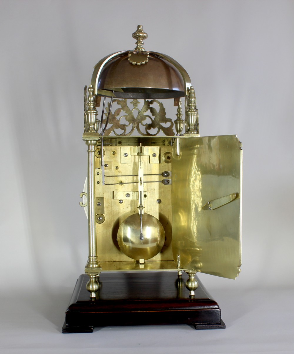 A brass twin fusee lantern clock featuring an engraved dial signed John Cator Woodbridge, a silvered Roman numeral chapter ring, a top bell structure, and mounted on a dark wooden base. View V