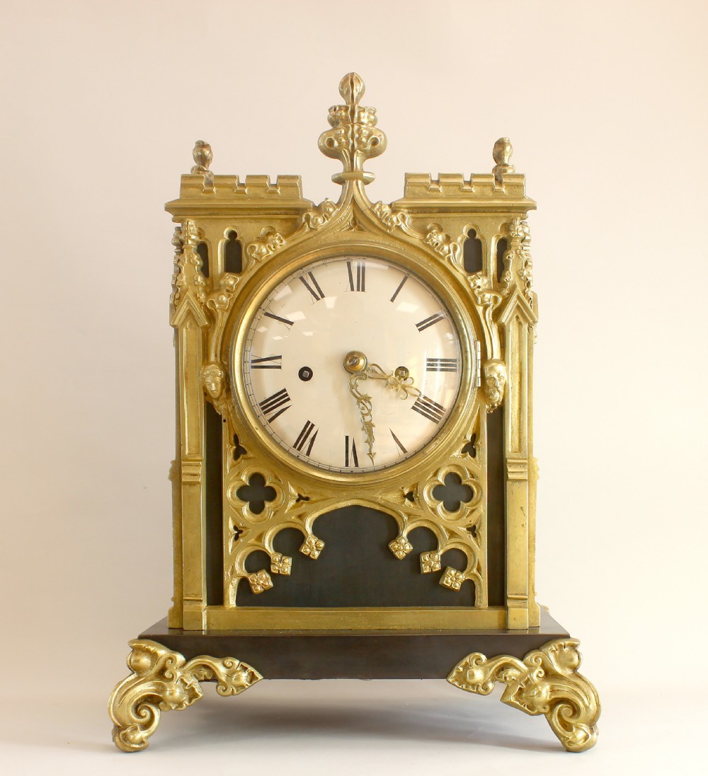 A Gothic Revival gilt brass and bronze mantel clock featuring architectural tracery, pointed arches, a white enamel dial with Roman numerals, and scroll feet.