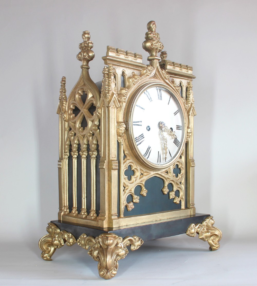 A Gothic Revival gilt brass and bronze mantel clock featuring architectural tracery, pointed arches, a white enamel dial with Roman numerals, and scroll feet. View III