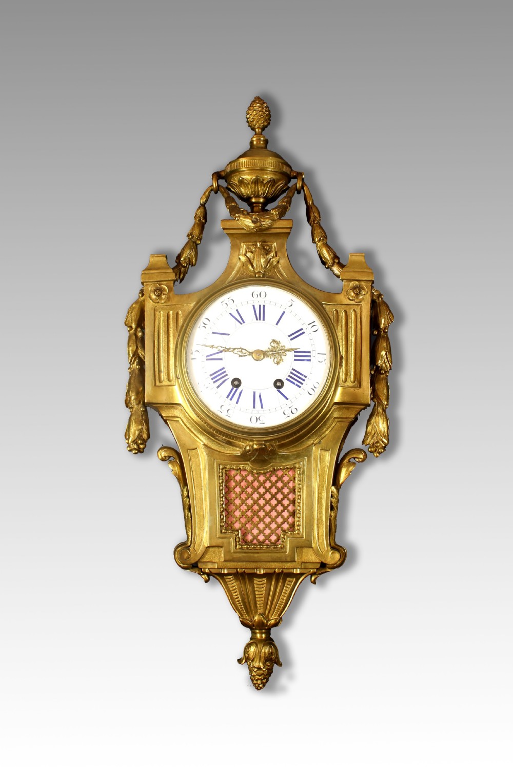 An ornate French gilt bronze cartel wall clock topped with a classical urn finial and decorated with sculpted garlands and a pink lattice panel.