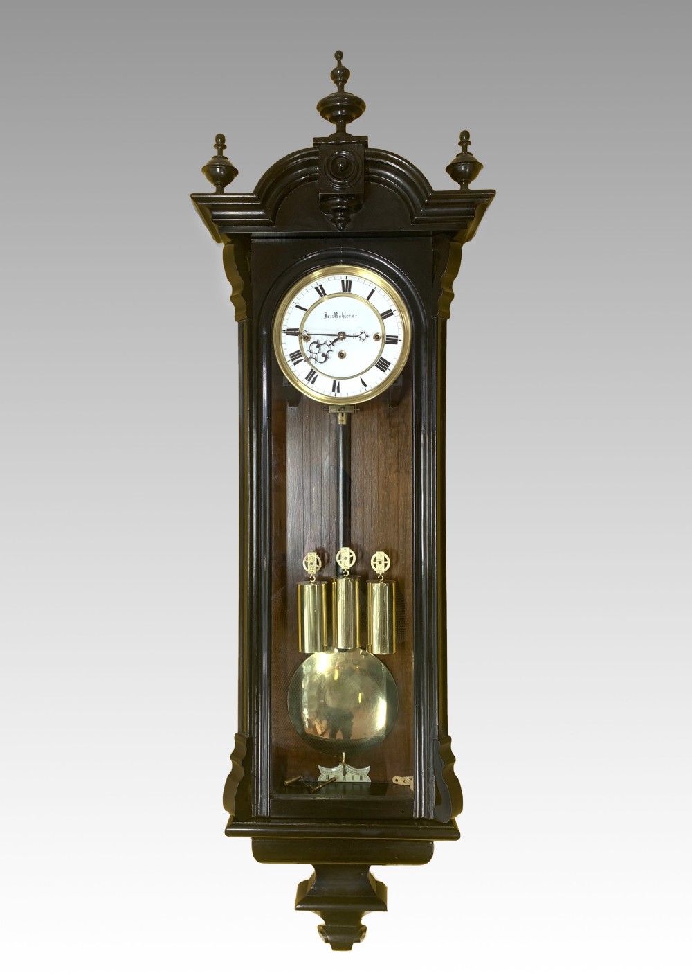 An ebonised wooden Vienna regulator wall clock featuring a glazed door, white enamel dial with Roman numerals, three brass weights, and a round brass pendulum bob.