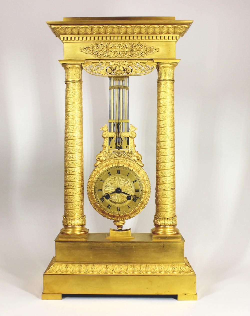 An antique French Empire style gilt bronze portico mystery clock featuring detailed columns supporting a decorated top frieze and suspending a circular dial with a gridiron pendulum.