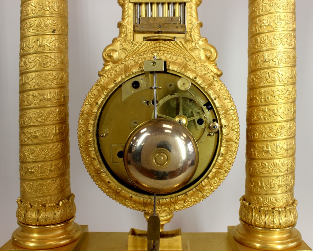 An antique French Empire style gilt bronze portico mystery clock featuring detailed columns supporting a decorated top frieze and suspending a circular dial with a gridiron pendulum. View VI