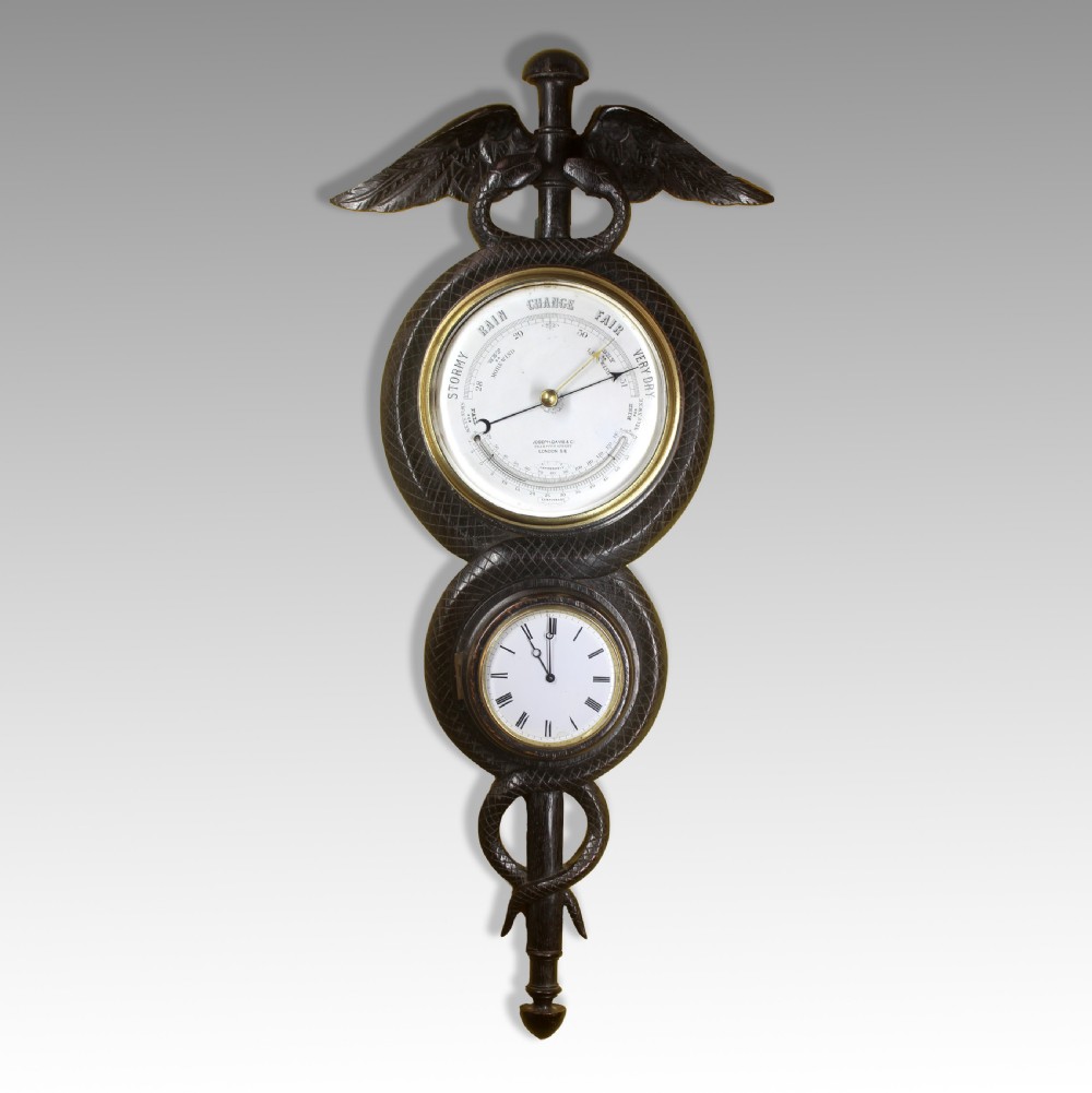 A dark carved wood wall-mounted clock and barometer combination sculpted in the shape of a caduceus staff with entwined snakes and wings.