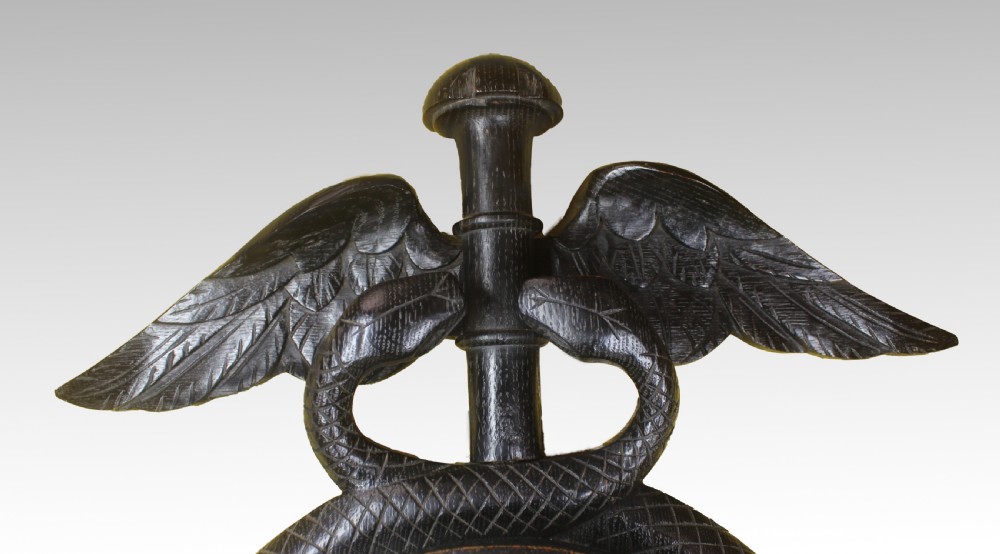 A dark carved wood wall-mounted clock and barometer combination sculpted in the shape of a caduceus staff with entwined snakes and wings. View II