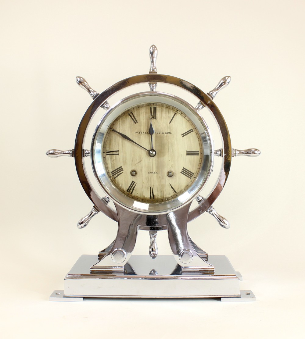 A chrome-plated nautical desk clock styled as a ship wheel mounted on a rectangular plinth with a cream dial and Roman numerals.