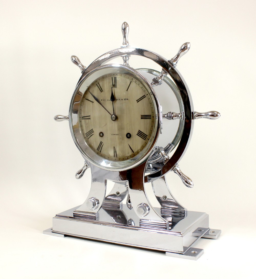 A chrome-plated nautical desk clock styled as a ship wheel mounted on a rectangular plinth with a cream dial and Roman numerals. View II