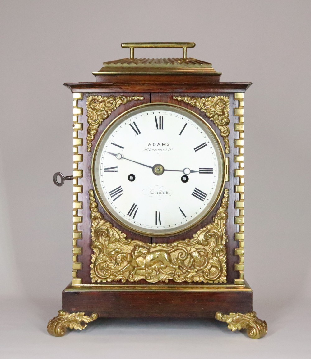 A mahogany and brass bracket clock featuring a white enamel dial with Roman numerals, gilt brass corner mounts, and scroll feet.